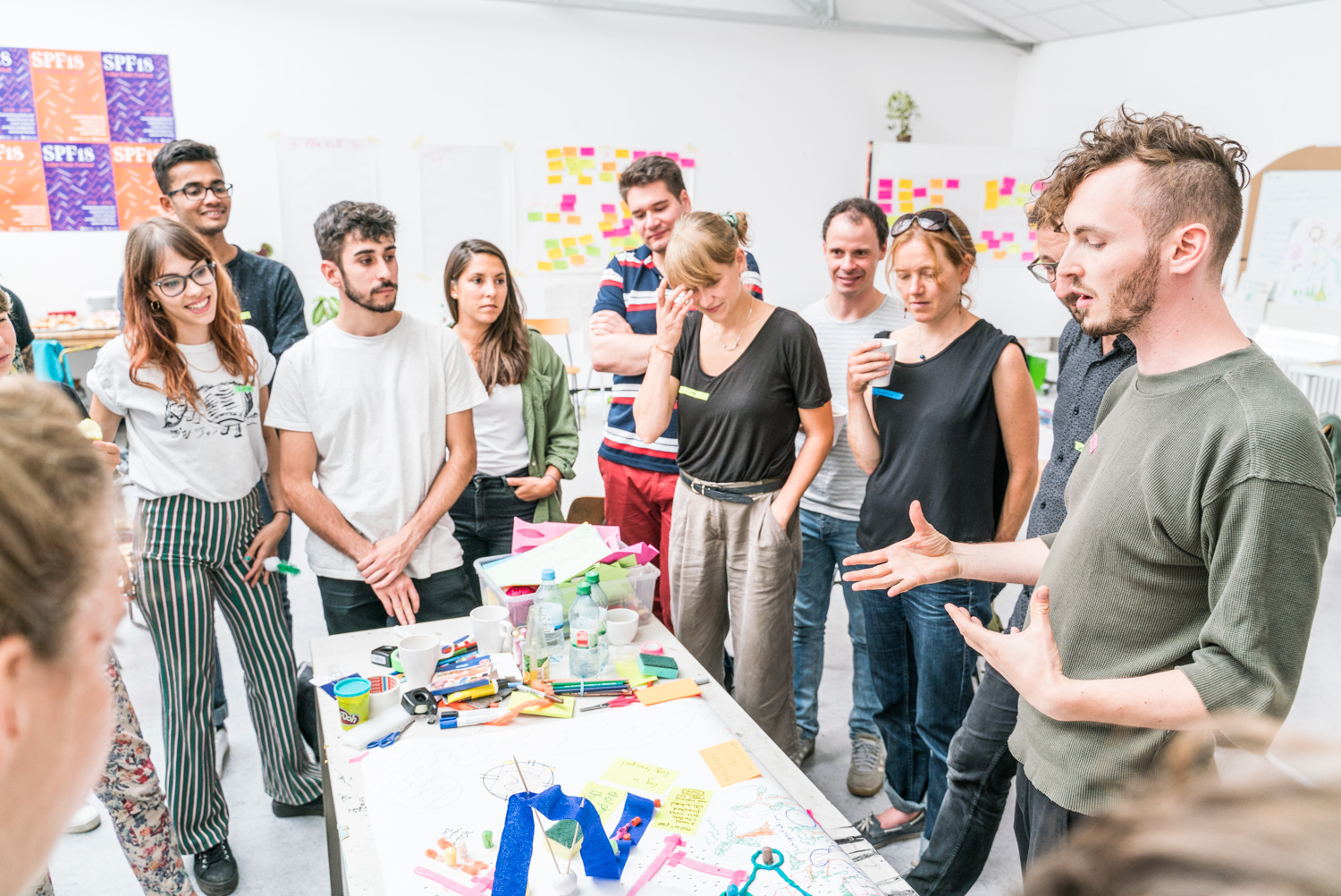 Workshop participants stand around a table with craft materials, discussing with a facilitator