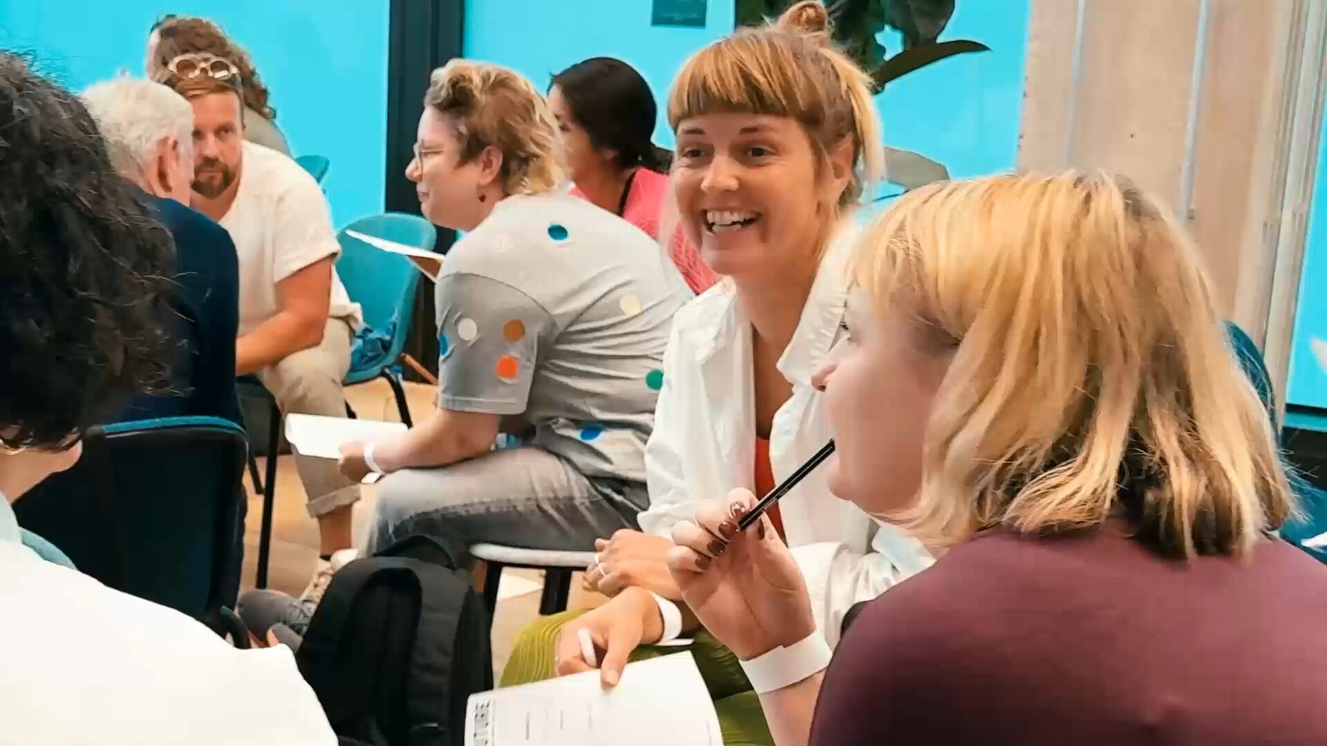 Teilnehmende sitzen in einer Gesprächsrunde zusammen, eine Frau lacht, im Vordergrund hält eine andere einen Stift